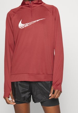 Persona que lleva una camiseta deportiva roja de manga larga con cremallera en el cuello y logo blanco de Nike, junto con pantalones cortos deportivos negros.