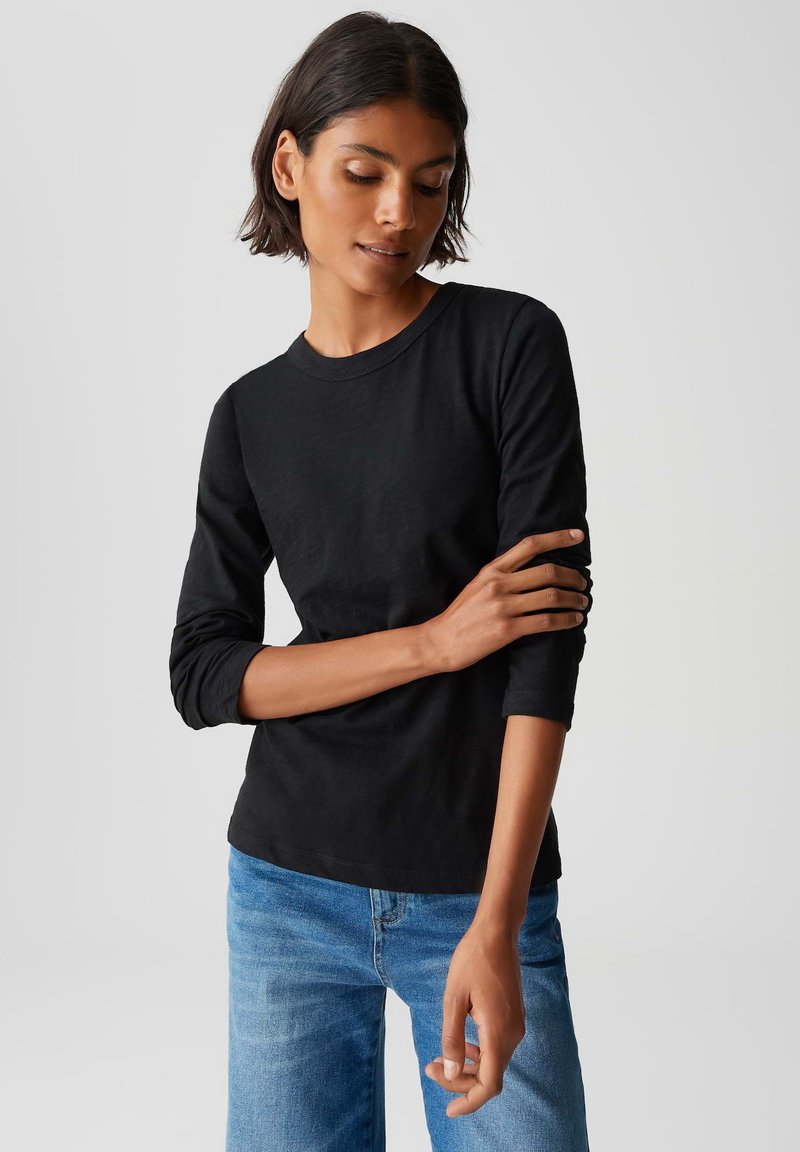 Frau mit kurzem dunklem Haar, die ein schwarzes Langarmshirt und blaue Jeans trägt und auf einem einfarbigen hellgrauen Hintergrund den Ärmel anpasst.