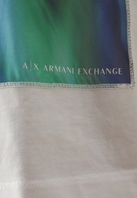 Tejido blanco que presenta un parche de degradado en verde y azul con el logo "A|X ARMANI EXCHANGE". El parche tiene una textura suave y un borde cosido.