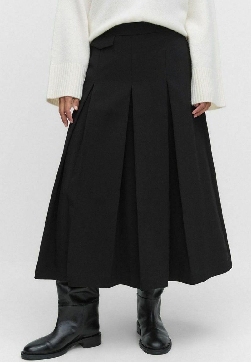 Sort plisseret midi-nederdel iført en hvid sweater og sorte læderstøvler til midt på læggen, vist fra talje til fødder.