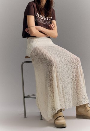 Personne assise sur un tabouret portant un t-shirt marron court avec du texte, une longue jupe blanche en dentelle et des baskets montantes beiges.