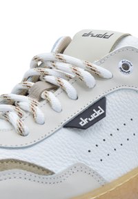 Ripresa ravvicinata di una sneaker in pelle bianca con accenti beige, lacci bianchi a motivo, perforazioni e il marchio "drudd" sulla linguetta e sul lato.