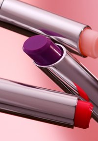 Tre läppprodukter i silvermetalliska tuber med rundade spetsar, i nyanser av lila, röd och rosa mot en mjuk rosa bakgrund.