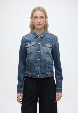 Kobieta o długich blond włosach, ubrana w dopasowaną niebieską kurtkę jeansową z zapięciem na guziki z przodu i kieszeniami na piersi, połączoną z czarnymi spodniami, na białym tle.