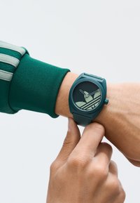 Mörkgrön silikonarmbandsur med en rund urtavla, med ljusgröna tim- och minutvisare och ett randigt mönster på urtavlan.