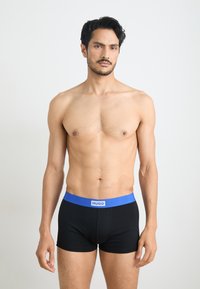 Black boxer briefs with a blue elastic waistband labeled "HUGO." Fabric is smooth, fitted design, worn above the hips on a bare torso.