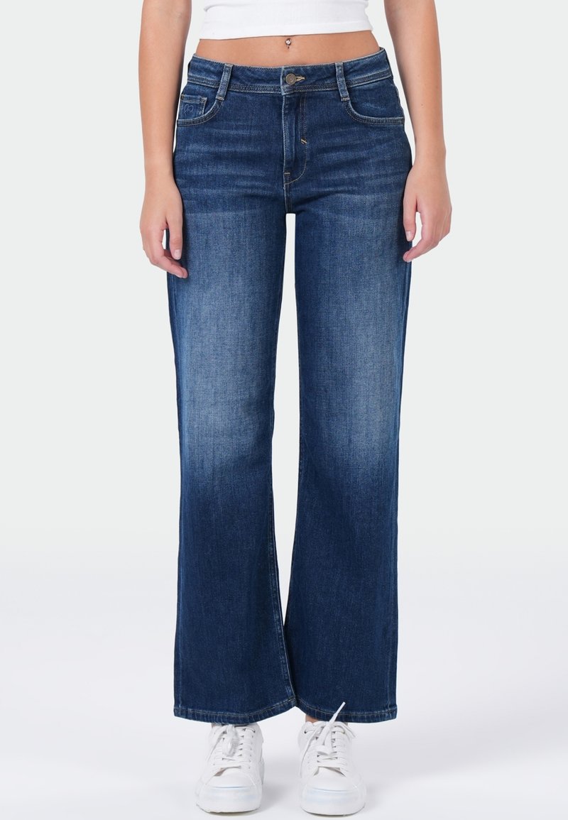 Vrouw in een mid-rise, wijd-uitlopende blauwe jeans met subtiele vervaging, gecombineerd met witte sneakers en een cropped wit topje.