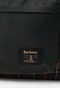 Dark green fabric with Barbour logo patch featuring a lighthouse and waves above a tartan fabric base.