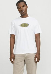 Tricou alb din bumbac cu mâneci scurte, având un logo central într-un design oval aurie și albastră, purtat cu pantaloni bej deschis.