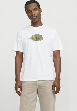 Hvid bomulds t-shirt med korte ærmer, som har et centralt logo i guld og blåt ovalt design, båret med lyse beige bukser.