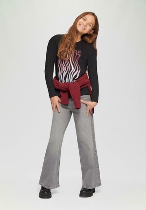 Zwart shirt met lange mouwen en abstracte grafiek, grijze flared jeans en een rode trui om de taille geknoopt; draagt zwart chunky schoenen.