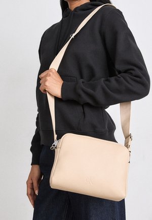 Person wearing black hoodie and dark pants, carrying a beige crossbody bag with an adjustable strap over the shoulder.