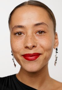 Närbild av ett ansikte med framträdande fräknar. Har djärva röda läppar och svarta abstrakta hängande örhängen. Slät hy och rakt mörkt hår.