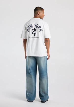 Mann trägt weißes T-Shirt mit "New York Club House" Text und Symbol auf dem Rücken, kombiniert mit weiten blauen Jeans, stehend vor einfachem Hintergrund.