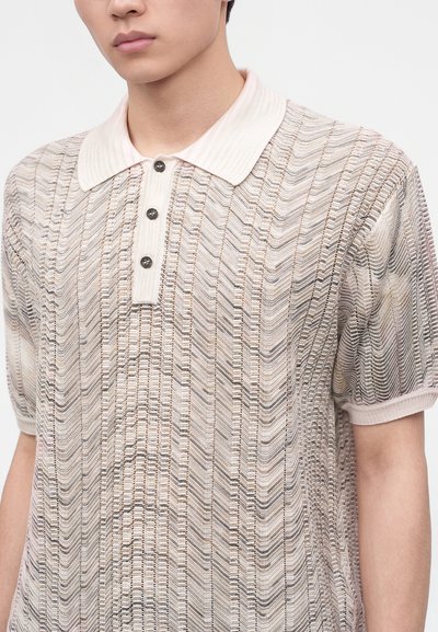 Jeune personne portant un polo texturé beige et blanc à manches courtes avec col en côte et trois boutons foncés, sur un fond clair uni.