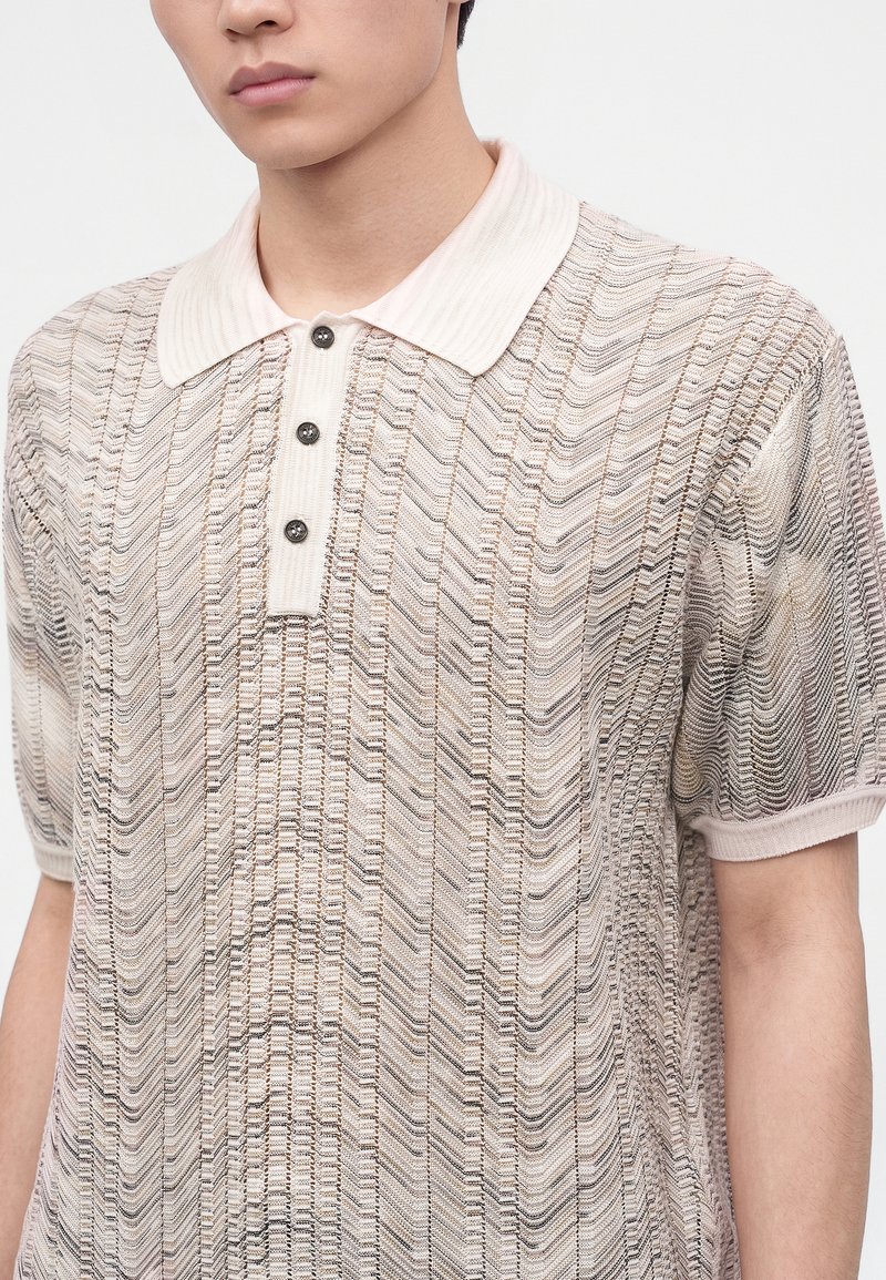 Persona joven con polo de manga corta texturizado en beige y blanco con cuello acanalado y tres botones oscuros, sobre un fondo claro y liso.