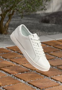 Bijeli kožni tenisice s glatkom teksturom, okruglim vrhom i ravnim vezicama. Imaju suptilan logo sa strane i elegantan minimalistički dizajn.