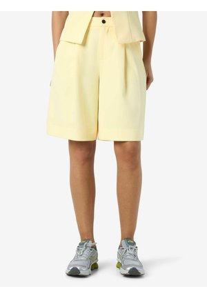 Personne portant un short sur mesure jaune pâle jusqu'aux genoux avec des plis, associé à des chaussures de course argentées et jaunes, montré de la taille aux pieds.