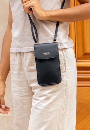 Black leather crossbody phone case with a flap closure, featuring a gold logo. The adjustable strap hangs from the user's shoulder.