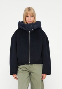 Mujer vestida con una chaqueta negra de gran tamaño con cremallera y un gran cuello acolchado, y pantalones verde claro, de pie frente a un fondo blanco liso.