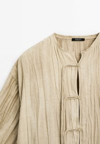 Haut beige à manches longues en tissu texturé, avec un col rond, une fermeture à boutons sur le devant et des plis verticaux.