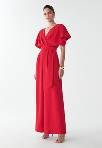 Punane, laia säärega kombekas lühikeste varrukatega, V-kaelusega ja sidumisega taljepiiril. Valmistatud sujuvast kangast. Kaunistatud kuldsete kõrvarõngaste ja rihmadega kontsadega.