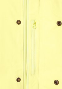 Gul jacka med en huvuddragkedja och en liten dragkedjeficka, bronzfärgade tryckknappar och en mjuk textur.