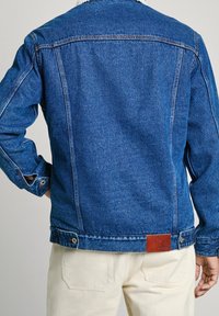 Denimjacka i mörkblått med en texturerad yta, med en shearlingkrage, knappmanchetter och en brun läderpatch vid midjan.