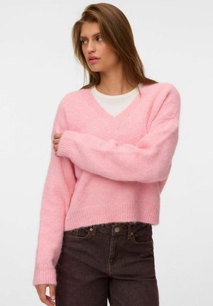 Femme regardant sur le côté portant un pull rose doux à col en V par-dessus un haut blanc et un jean marron foncé sur un fond blanc uni.