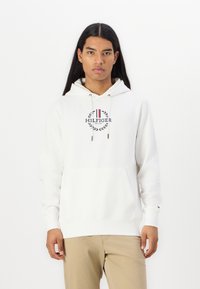 Hvit hettegenser med frontlomme, med et sirkulært logo i marineblått og rødt, og snorer med metallspisser.