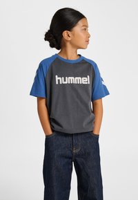 Grå och blå kortärmad t-shirt med texten "hummel", med raglan-design och mjuk bomullstextur, kombinerad med mörka denimjeans.