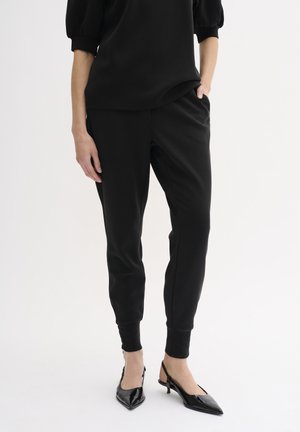Jogger neri in cotone misto con polsini elastici, dotati di tasche laterali, abbinati a scarpe nere in vernice con punta a ogiva. Texture liscia in tutto il capo.