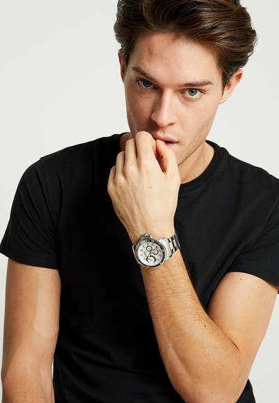 Une montre en acier inoxydable argenté avec un cadran rond, des accents noirs et un affichage de la date, portée au poignet, avec un t-shirt noir visible.