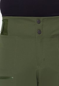 Pantalons taille haute vert olive avec deux boutons en métal noir, texture lisse, poches latérales et ceinture large avec un accent en tissu.
