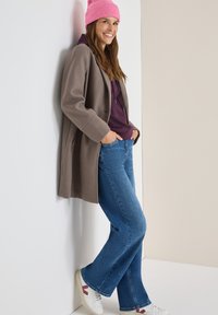 Abrigo de lana gris sobre una sudadera morada, combinado con jeans acampanados azules y zapatillas blancas con acentos rojos. Gorro de punto rosa en la cabeza.