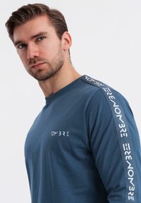 Mladý muž s upraveným vousem, oblečený v modré košili s dlouhým rukávem, na které je na hrudi a na ramenech nápis "OMBRE".