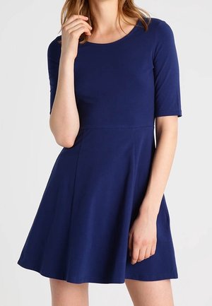 Vrouw draagt een knielange, donkerblauwe jurk met korte mouwen, staand met één hand bij haar gezicht en de andere ontspannen langs haar zijde.