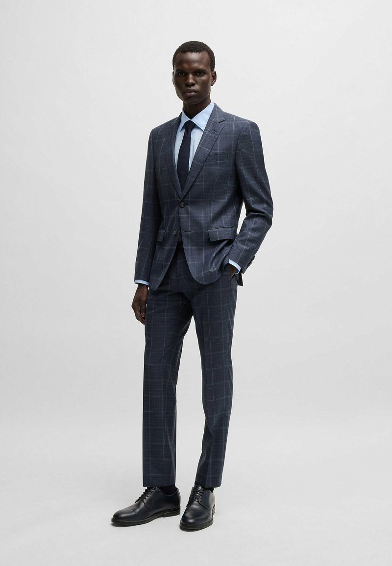 Abito a quadri blu navy con vestibilità su misura, abbinato a una camicia azzurra chiara e una cravatta scura. Scarpe in pelle nera completano il look.