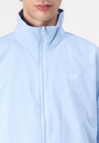 Ljusblå zip-jacka med texturerad yta och hög krage. Utrustad med en broderad logotyp på vänster sida. Hållbart material.