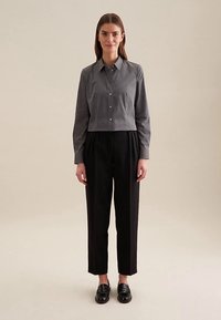 Graues Hemd mit Knopfleiste, Kragen, kurz geschnitten, und schwarze plissierte Hose. Die Schuhe sind schwarze Loafers mit glatter Oberfläche und minimalen Details.