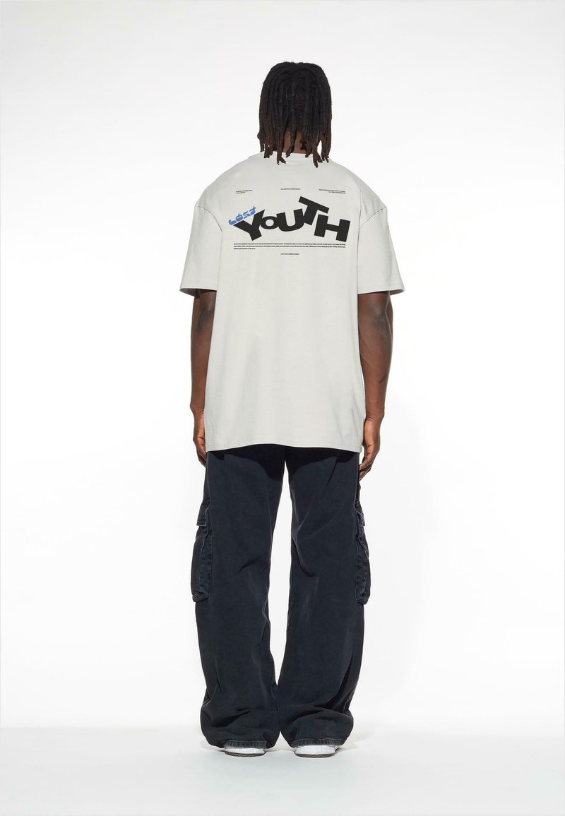 Šviesiai pilka didelio dydžio marškinė su "Jaunimo" grafika juodu ir mėlynu tekstu nugaroje, derinami su tamsiomis kargo kelnėmis.