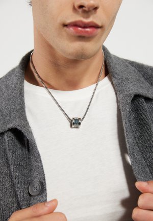 Metal necklace with a cylindrical pendant featuring silver and blue accents, worn over a white shirt and gray cardigan. Soft texture.