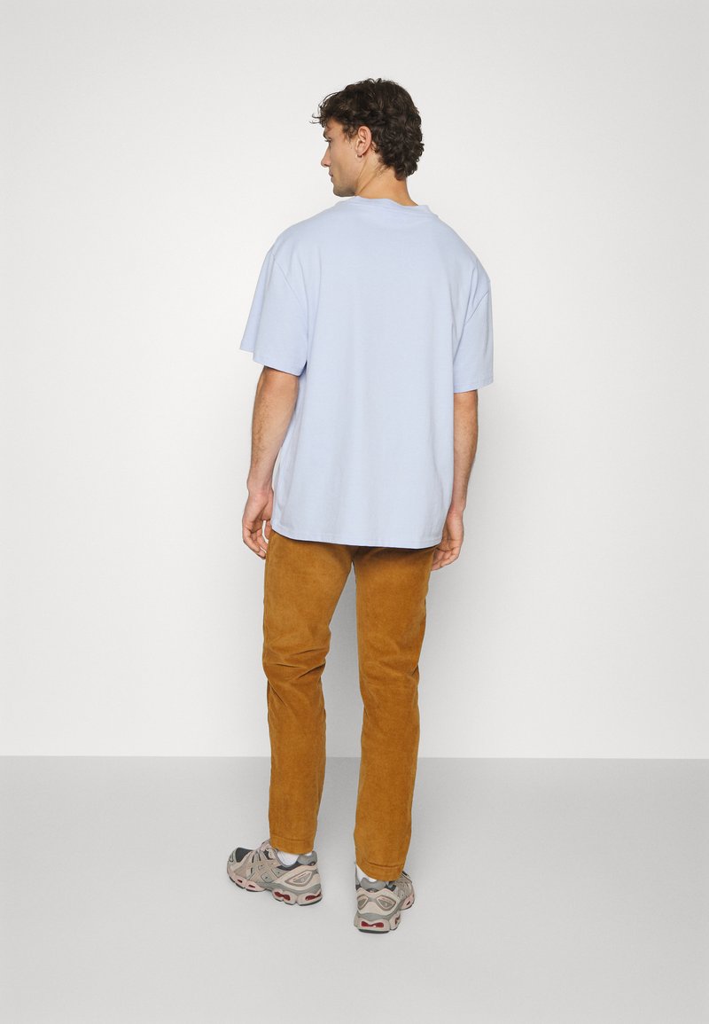 Homme debout, de dos, portant un T-shirt oversized bleu clair, un pantalon en velours côtelé marron et des chaussures de sport grises sur un fond uni.