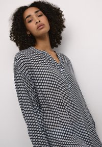 Blusa a pois in bianco e nero con scollo a V e bottoni, caratterizzata da una vestibilità larga e un tessuto strutturato. Maniche lunghe con un leggero drappeggio.