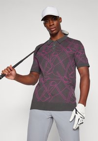 Gray polo shirt with pink golf club pattern, featuring a buttoned collar, short sleeves, and ribbed hem. Model holds a golf club.