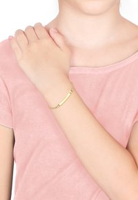 Guldarmband med en slät, förlängd stav och kedjelänkar. Bärs på handleden, kompletterad med en mjuk rosa skjorta.