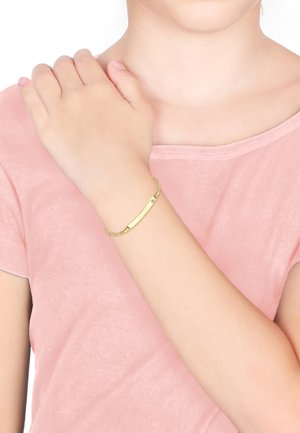 Goldarmband mit einem glatten, länglichen Balken und Kettenverbindungen. Wird am Handgelenk getragen und ergänzt von einem sanften rosa Hemd.