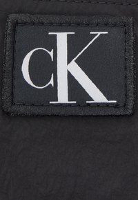 Černá látková etiketa má v tučném bílém písmu nápis "ck". Okolní šití je černé a etiketa má texturovaný povrch.