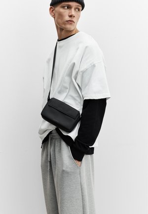 Ung mand iført lagdelte sort-hvide toppe med grå joggingbukser, iført en sort beanie og en sort crossbody taske, ser mod højre.