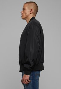Veste bomber noire avec une texture lisse et légèrement brillante ; poignets et taille côtelés ; comprend une poche latérale et une coupe décontractée.
